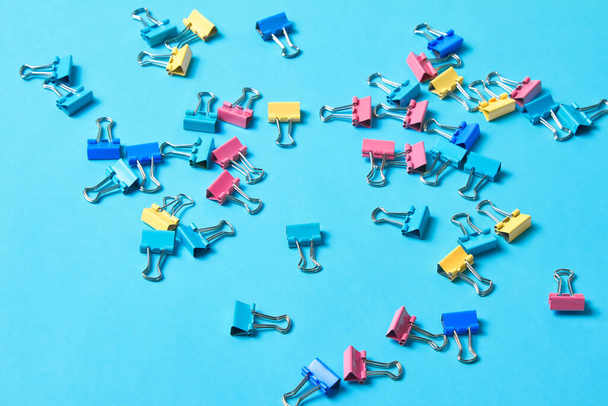 Lot of cute colorful office binder clips laying on turquoise background in disorder.Some empty space left between.Side view of chaotic position of blue,pink,yellow paper holders on surface.Horizontal - Foto, Bild
