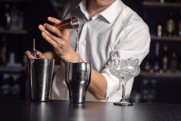 Cropped picture of hand of bartender dressed in white shirt. Glass vase with ice cubes on right side.Cocktail equipment on black shiny surface in focus.Horizontal layout - Photo, Image