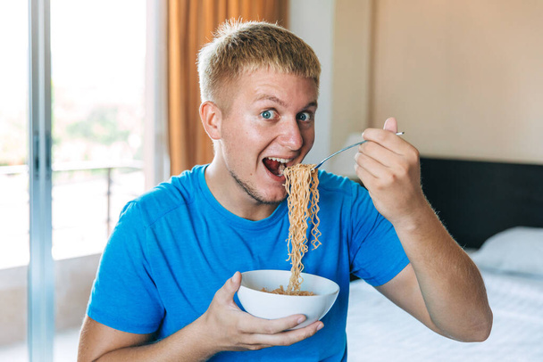 Young handsome man in a blue T-shirt eating instant noodles from a white plate for lunch at home, concept of cheap food or food delivery, selective focus, close-up - Photo, Image