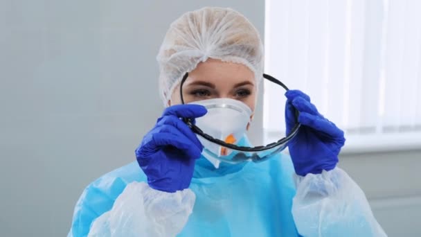 Female doctor in a protective uniform, gloves and respirator is preparing to receive patients. She puts on a glasses. The camera moves sideways and approaches the face. Woman looking at camera - Séquence, vidéo