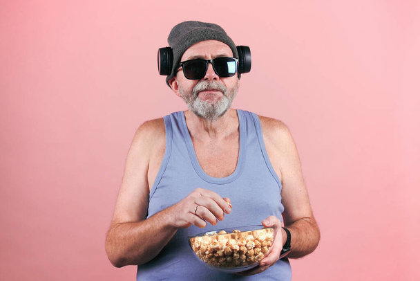 Menő, érett férfi kék felsőben, napszemüveg, szürke kalap, vezeték nélküli fejhallgató és egy tál popcorn a szájba dobva. Rózsaszín alapon izolált. - Fotó, kép