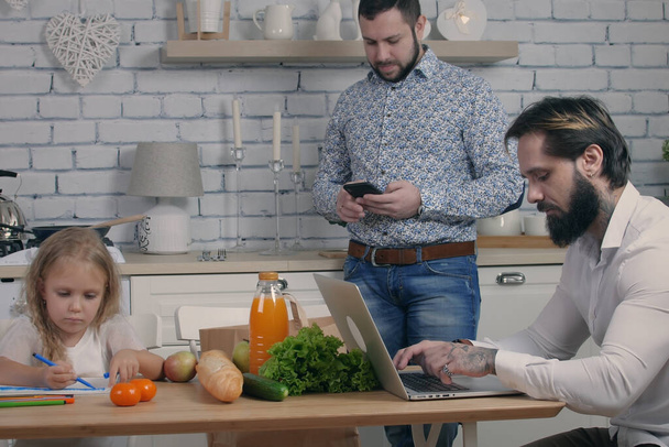 Férfi pár a lányukkal a konyhában férfiak segítségével kütyü laptop és mobiltelefon kis szőke lány rajz az asztalnál, egyenlőség modern család otthon - Fotó, kép