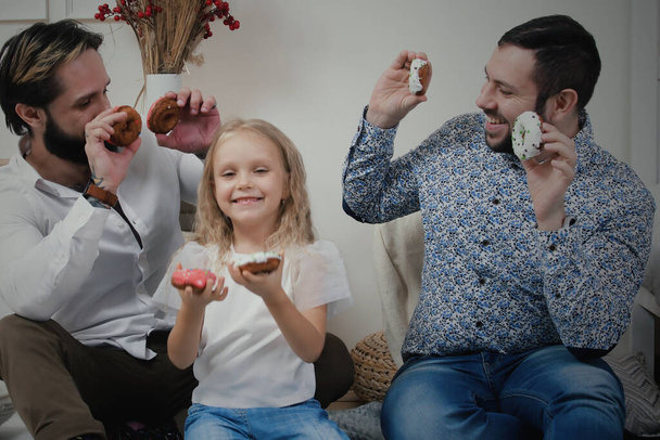 İki şık genç adam ve beş yaşında bir kız çocuğu dairede yerde oturup jöleli çöreklerle oynuyorlar. Homoseksüel erkek ve kızları donutlarla komik suratlar yapıyor. - Fotoğraf, Görsel