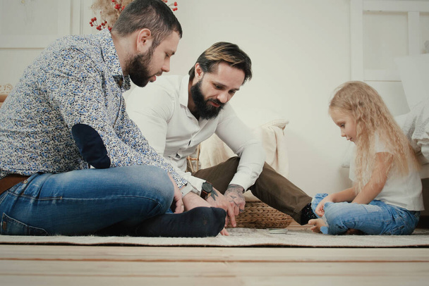 Das männliche Paar und seine fünfjährige Tochter spielen ein Brettspiel, während sie in der Wohnung auf dem Boden sitzen. Sie haben Spaß und lachen - Foto, Bild