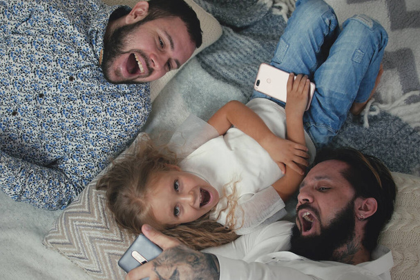 Top View Homoseksuaalinen pari ja heidän viisivuotias tytär ottaa hauskaa ottaa selfietä älypuhelimella kamera App tehdessään kasvoja ja huutaa. Samaa sukupuolta pari tyttären kanssa hauskaa
 - Valokuva, kuva