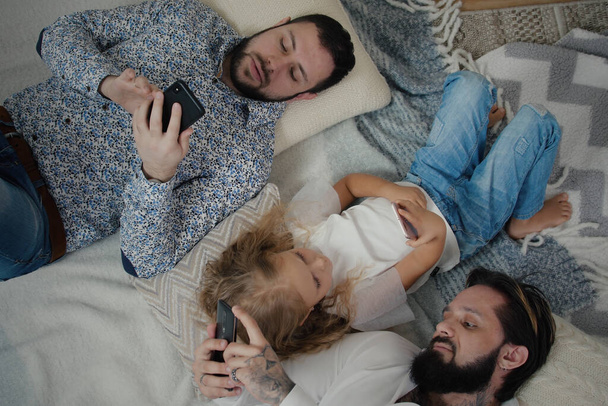 Πάνω άποψη των δύο κομψών ανδρών και μικρό χαριτωμένο κορίτσι βρίσκεται στο πάτωμα και να δούμε τα τηλέφωνά τους. Ίδιο ζευγάρι σεξ με την κόρη ξαπλωμένη στο πάτωμα στο σπίτι κάνοντας σερφ στα μέσα κοινωνικής δικτύωσης ή στο διαδίκτυο - Φωτογραφία, εικόνα