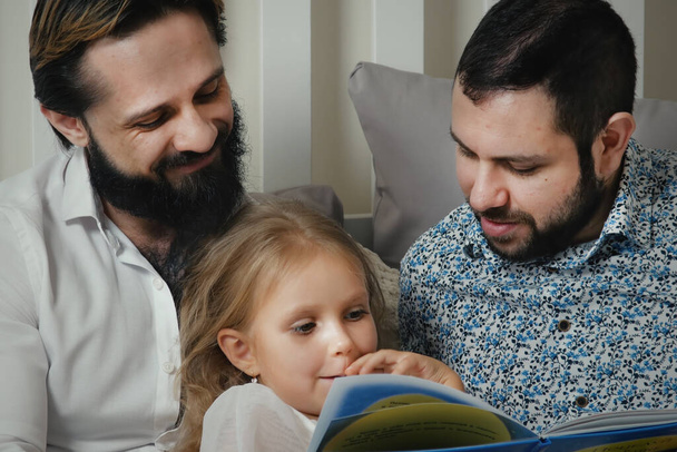 Aynı cinsiyetten erkek çift yatakta kızlarıyla yatıp kitap okuyor. Çocuklu şık eşcinsel çift. - Fotoğraf, Görsel