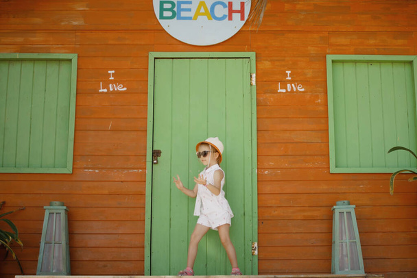5 letá běloška v klobouku a slunečních brýlích smyslně tančí hladce na pozadí oranžového plážového domu - Fotografie, Obrázek