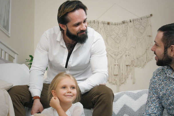Same Sex Male Couple At Home. ombing daughters hair and talking with a partner while sitting in bedroom. Stylish gay couple with their child - Photo, Image
