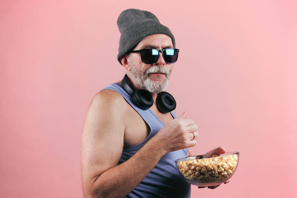 Πλάτη άποψη πλάνα αρχείου ενός ηλικιωμένου άνδρα σε μπλε μπλουζάκι, γυαλιά ηλίου, γκρι καπέλο και ασύρματα ακουστικά γύρω από το λαιμό στροφή μισό στην κάμερα με μπολ ποπ κορν. Απομόνωση σε ροζ φόντο. - Φωτογραφία, εικόνα
