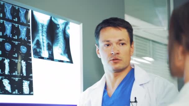Doctor male is showing the problem area on the x-ray photograph to the woman. Spinal tomography is hanging on the white board and the doctor discussing the results. - Filmagem, Vídeo