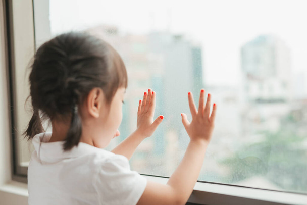 小さなアジアの女の子子供が家に座って窓を見て. - 写真・画像