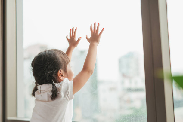Πίσω όψη σε χαριτωμένο κοριτσάκι κοιτάζει έξω από το παράθυρο - Φωτογραφία, εικόνα
