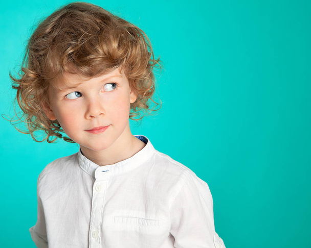 A beautiful curly-haired, blue-eyed boy looks slyly, incredulously, mysteriously to the side. In a white shirt against a sea-green background. Portrait of a beautiful four-year-old child. - Foto, afbeelding