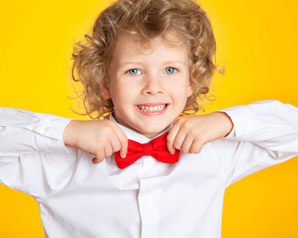 Curly-haired boy with blond hair and blue eyes in a shirt and a red bow tie on a yellow background. The child is laughing, smiling, having fun, happy. - Foto, afbeelding