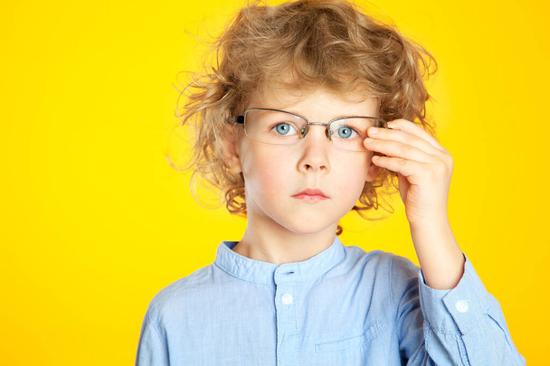 A beautiful European curly-haired boy with glasses looks thoughtfully, seriously, adjusts the frames of his glasses. A child in a shirt on a yellow background. A botanist, a Junior high student. - Fotografie, Obrázek