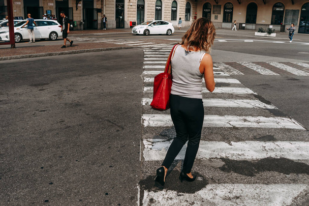 A woman with curly hair and a red shoulder bag crosses an intersection on a pedestrian crosswalk | BOLOGNA, ITALY - 17 SEPTEMBER 2018. - Zdjęcie, obraz