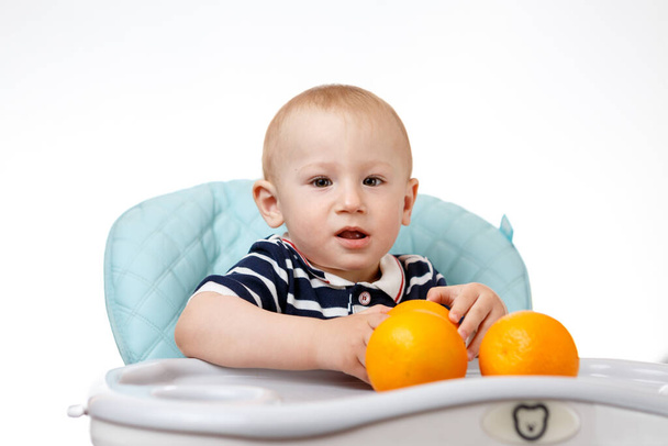 A cheerful boy is sitting in a baby seat and eating oranges on a white background. Fresh fruits are very beneficial for children. Smart adults are good nutritionists. - 写真・画像
