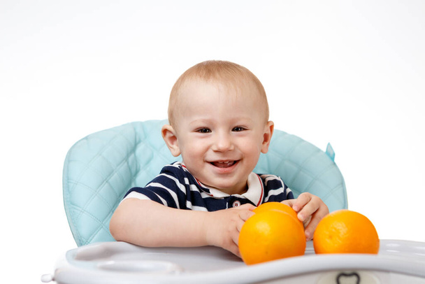 A cheerful boy is sitting in a baby seat and eating oranges on a white background. Fresh fruits are very beneficial for children. Smart adults are good nutritionists. - Foto, Imagem
