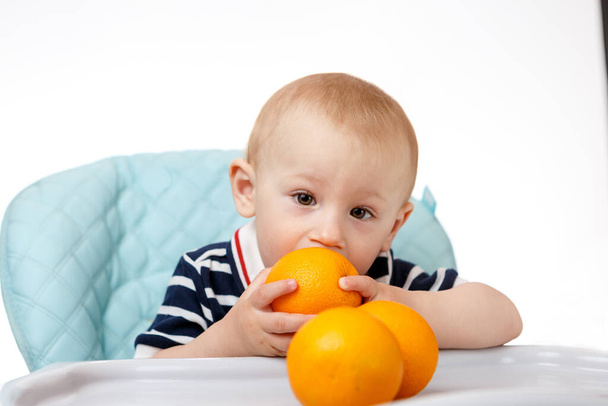 A cheerful boy is sitting in a baby seat and eating oranges on a white background. Fresh fruits are very beneficial for children. Smart adults are good nutritionists. - Foto, afbeelding