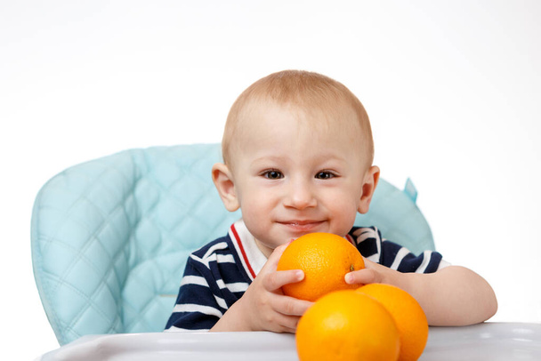 A cheerful boy is sitting in a baby seat and eating oranges on a white background. Fresh fruits are very beneficial for children. Smart adults are good nutritionists. - Valokuva, kuva