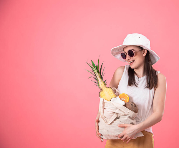 Portrét mladé stylové ženy oblečené v letních šatech s kloboukem a slunečními brýlemi, držící tašku s ECO ovocem, na růžovém izolovaném pozadí. - Fotografie, Obrázek