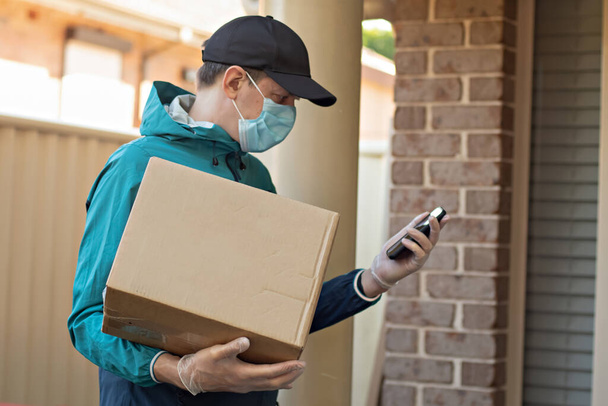 Contactless delivery during COVID-19 pandemic lockdown concept. Courier wearing mask and gloves holds a parcel box and checking address details on the smartphone - Zdjęcie, obraz