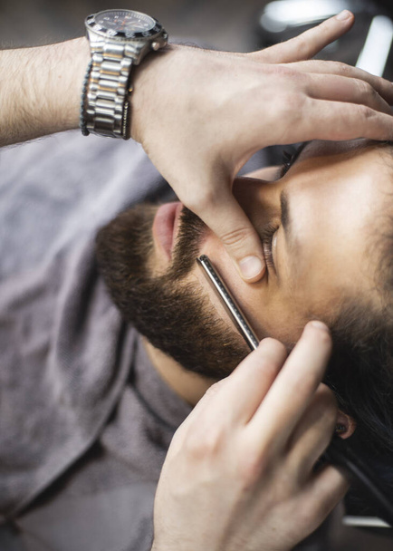 Barber hands with a razor shaving beard of male customer - Фото, зображення