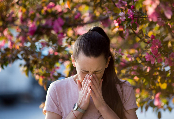 Young pretty woman sneezing in front of blooming tree. Spring allergy concept - Photo, Image
