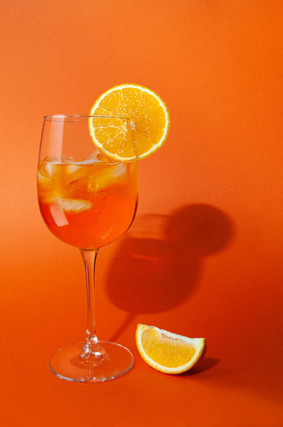 Cocktail aperol spritz with ice. In a glass goblet. On a glass of orange slice. Next to the glass is a slice of orange. On a plain orange background. Hard light, sharp shadows. Close-up. Place for text. - Photo, Image