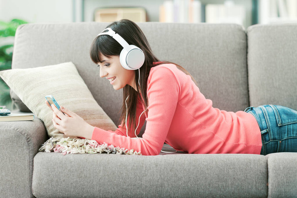 Young smiling woman with headphones relaxing on the sofa and listening to music online using a smartphone - Fotografie, Obrázek