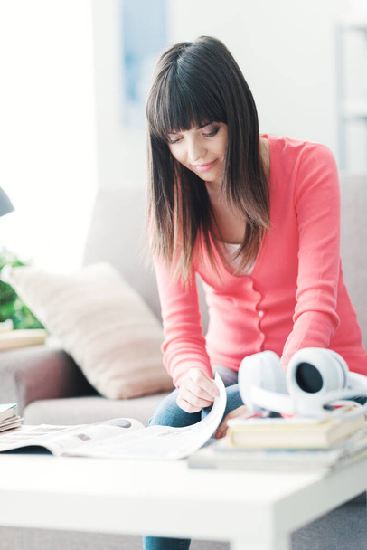 Ελκυστική νεαρή γυναίκα που χαλαρώνει στο σπίτι, κάθεται στον καναπέ και διαβάζει ένα περιοδικό - Φωτογραφία, εικόνα