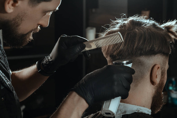 BARBERSHOP THEME. BEARDED BARBER IN BLACK RUBBER GLOVES IS TRIMMING THE HAIRCUT OF HIS YOUNG SERIOUS CLIENT. HE IS USING A HAIR CLIPPER - Fotografie, Obrázek