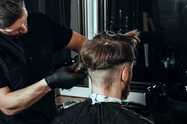 BARBERSHOP THEME. BEARDED SERIOUS BARBER IN BLACK RUBBER GLOVES IS TRIMMING THE HAIR OF HIS YOUNG HANDSOME CLIENT WITH CLOSED EYES. HE IS USING A HAIR CLIPPER - Fotografie, Obrázek