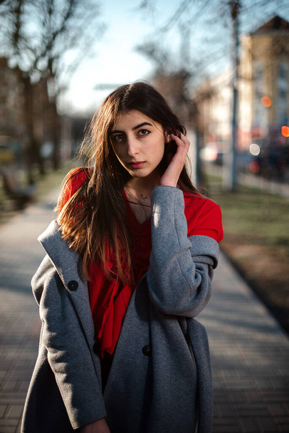 Girl in a red blouse and a gray cardigan on a blurry city background in early spring - Foto, afbeelding
