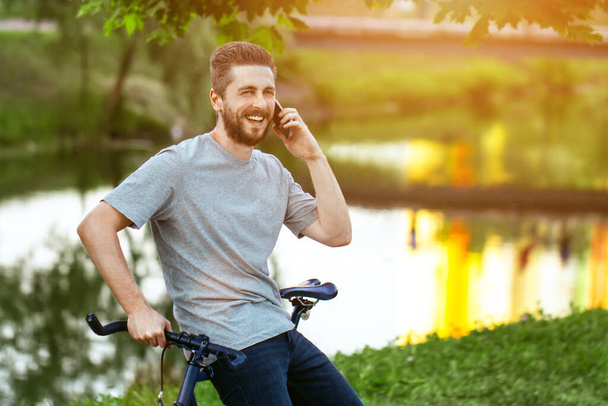 Handsome Young Man In Grey T-Shirt And Blue Jeans Stops Cycling, To Talk On The Phone In An Alley With Green Trees Near Lake - Foto, imagen
