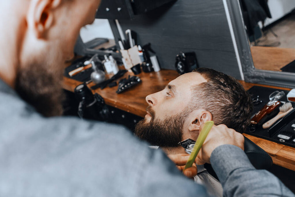BARBERSHOP THEME. BALD BEARDED BARBER IS TRIMMING THE BEARD OF HIS YOUNG HANDSOME CLIENT. HE IS USING A HAIR CLIPPER - Fotoğraf, Görsel