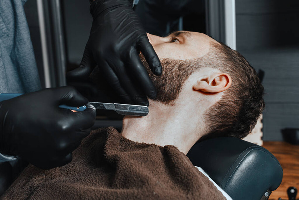 BARBERSHOP THEME. BARBER IN BLACK RUBBER GLOVES IS TRIMMING THE BEARD OF HIS YOUNG HANDSOME CLIENT. HE IS USING A  STRAIGHT RAZOR - Foto, Imagem