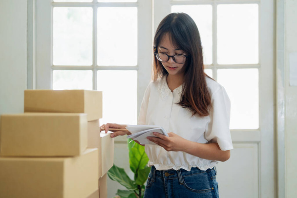 Shipping shopping online. Young entrepreneur working online e-commerce shopping at her shop. Young online seller checking order from customer and prepare parcel box deliver to customers. Online selling and online business e-commerce concept. - Photo, Image