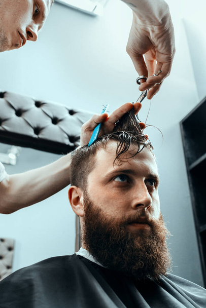 BARBERSHOP THEME. BARBER HOLDS A PAIR OF SCISSORS AND A COMB AND SHEARS THE CLIENT IN BARBER SHOP. BEARDED MAN SITTING IN BARBER CHAIR - Zdjęcie, obraz