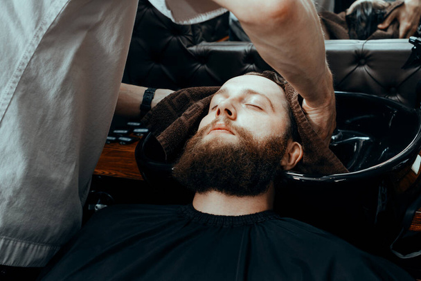 BARBERSHOP THEME. BARBER WIPING THE CLIENT'S HEAD AFTER WASHING IN BARBER SHOP. BEARDED MAN SITTING IN BARBER CHAIR - Foto, afbeelding