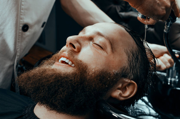 BARBERSHOP THEME. BARBER WASHING CLIENTS HAIR IN BARBER SHOP. SMILING BEARDED MAN SITTING IN BARBER CHAIR - Photo, image