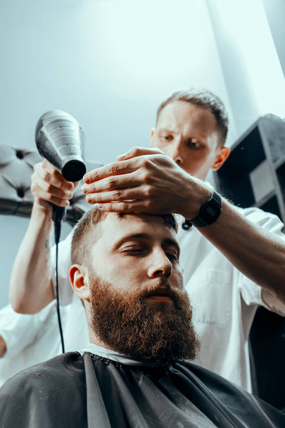 BARBERSHOP THEME. BARBER HOLDS A HAIR DRYER AND DRIES THE CLIENT'S HEAD AFTER WASHING IN BARBER SHOP. BEARDED MAN SITTING IN BARBER CHAIR  - Foto, imagen