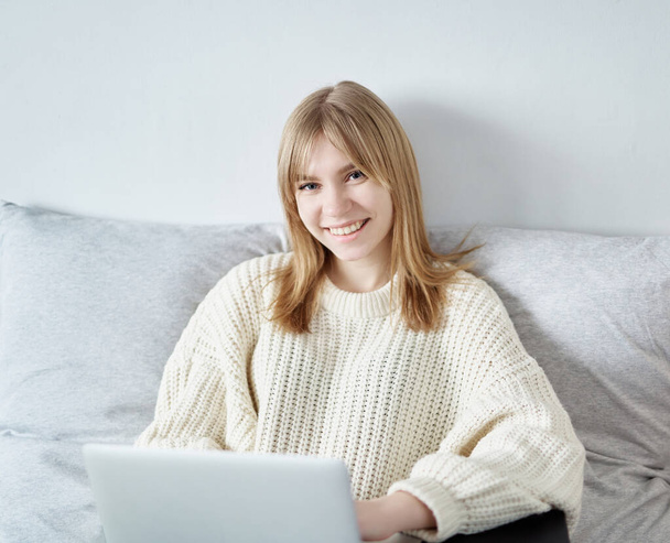 Atractiva joven sonriente mujer en suéter y jeans con portátil mirando a la cámara y sonriendo. Estudio en línea, trabajo, navegación, navegación por Internet inalámbrico en el hogar, concepto de tecnología
 - Foto, Imagen