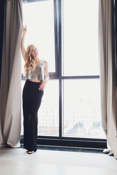 beautiful blonde woman in black clothes with heels stands by the window. rear view. - Foto, Imagem