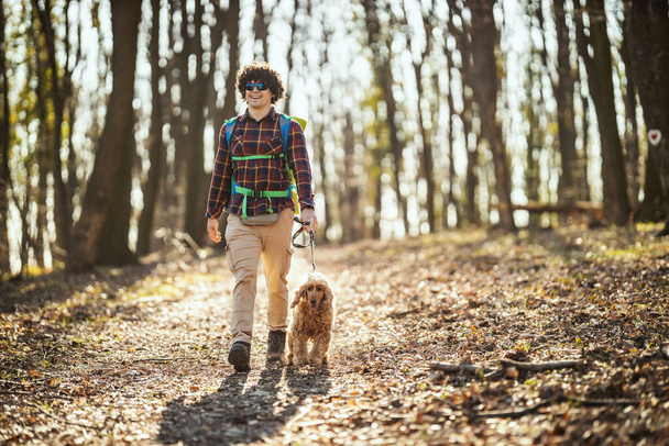 Happy smiling handsome man is walking along the forest path with his dear cute dog enjoying landscape in forest in beautiful sunny day. - Foto, imagen