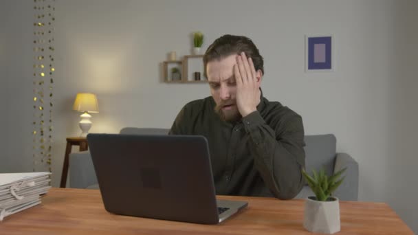 Ofis çalışanı sakallı bir adam dizüstü bilgisayarlı bir masaya oturur, bir laptop monitörüne bakar, bir adam yorgundur, evden uzakta çalışır. - Video, Çekim