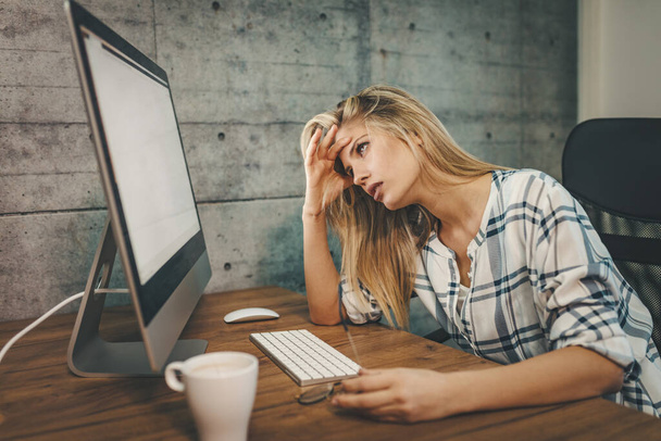 Mladá vyčerpaná a unavená návrhářka sedí v kanceláři před počítačem a drží hlavu, čeká na konci pracovní doby. - Fotografie, Obrázek