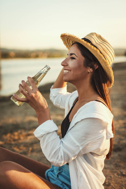 Молода жінка в солом'яному капелюсі відпочиває під час заходу сонця на березі річки і п'є пиво
. - Фото, зображення