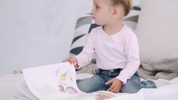Cute toddler girl is flipping a books pages on a bed - Séquence, vidéo